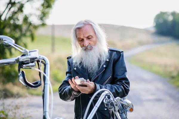 Senior man traveller z motocyklem na wsi, za pomocą smartfona. — Zdjęcie stockowe