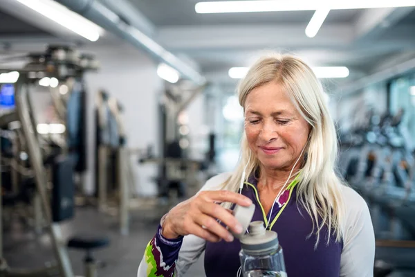 Una donna anziana con auricolari e bottiglia d'acqua in piedi in palestra . — Foto Stock