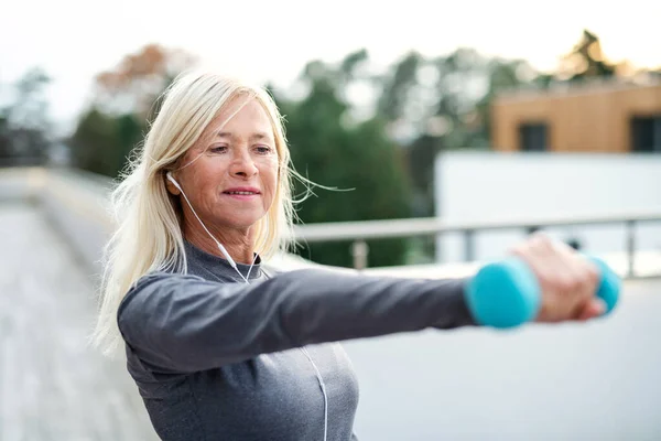 En äldre kvinna med hörlurar och hantlar utomhus som motionerar. — Stockfoto