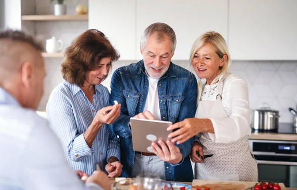 Group of senior friends with tablet at dinner party at home, cooking. — Stockfoto