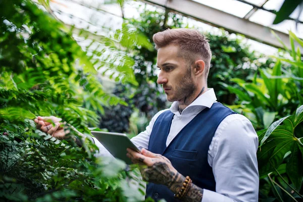 Young man with tablet standing in botanical garden, working. — Stockfoto
