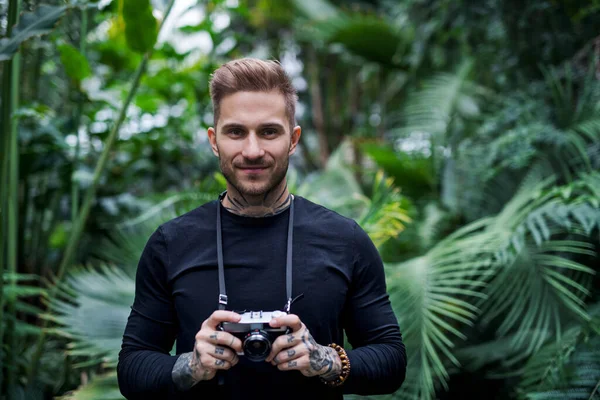 Front view of young man with camera standing in botanical garden. — Stock Photo, Image