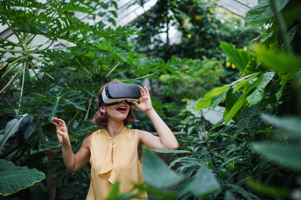 Vista frontal de la joven mujer de pie en el jardín botánico, usando gafas VR . — Foto de Stock