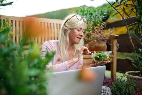 Mujer mayor con portátil sentado al aire libre en la terraza, almorzando . — Foto de Stock