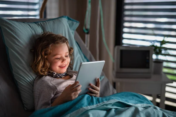 Uttråkad liten flicka i sängen på sjukhus, med hjälp av tablett för att fördriva tiden. — Stockfoto