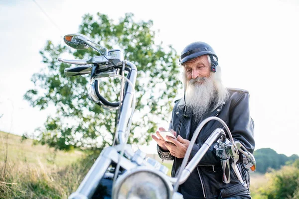Senior man traveller z motocyklem na wsi, za pomocą smartfona. — Zdjęcie stockowe