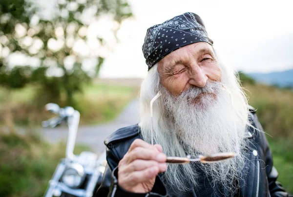 Senior man traveller with motorbike in countryside, winking. — Stockfoto