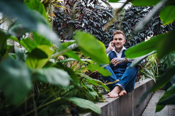 Young man with smartphone sitting in botanical garden, making phone call. — Stock Photo, Image