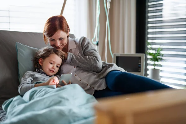 Liten flicka med mamma i sängen på sjukhus, med hjälp av smartphone för att fördriva tiden. — Stockfoto