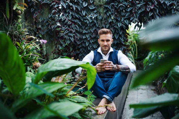 Young man sitting barefoot in botanical garden, using smartphone. — Stock Photo, Image