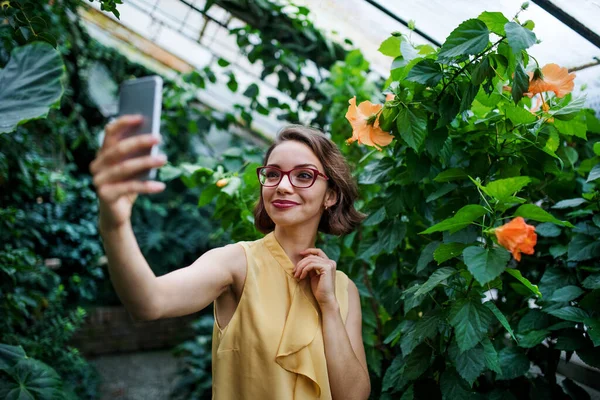 スマートフォンを持つ若い女性が植物園に立って、自撮り. — ストック写真