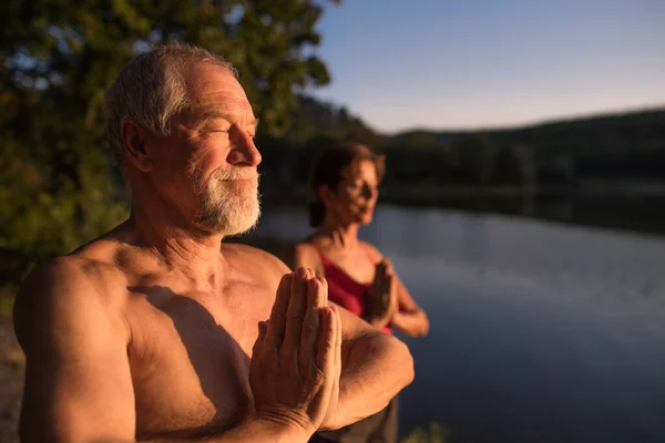 Старша пара в купальнику стоїть біля озера на відкритому повітрі в сутінках робить йогу . — стокове фото