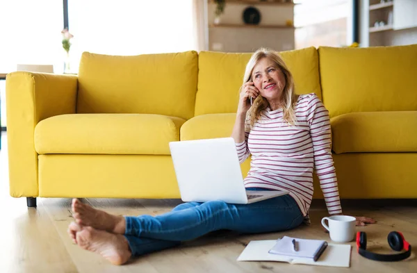 Senior vrouw met smartphone en laptop binnen in het thuiskantoor, werken. — Stockfoto