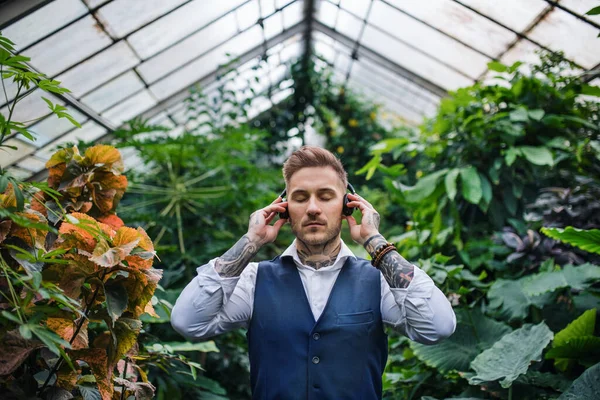 Young man with headphones standing in greenhouse in botanical garden. — Stock Photo, Image