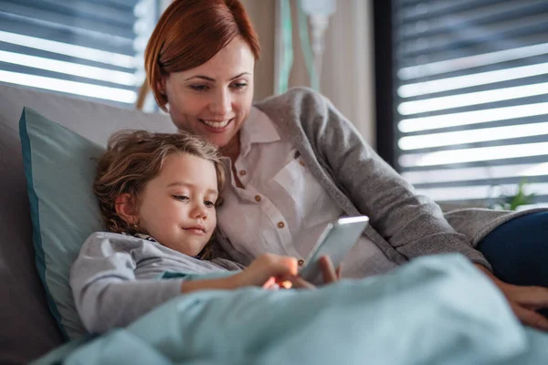 Niña pequeña con madre en la cama en el hospital, utilizando el teléfono inteligente para pasar el tiempo . — Foto de Stock