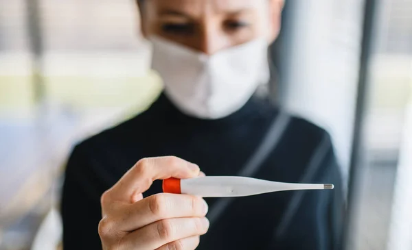 Femme inquiète avec thermomètre et masque facial à l'intérieur à la maison, concept de virus Corona . — Photo