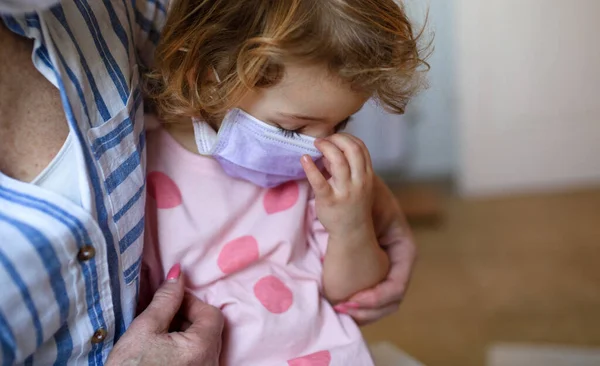 Grootmoeder en klein meisje met gezichtsmasker binnenshuis, corona virus en quarantaine concept. — Stockfoto