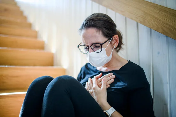 Vrouw met gezichtsmasker en pijn op de borst thuis zittend, Corona virus en quarantaine concept. — Stockfoto