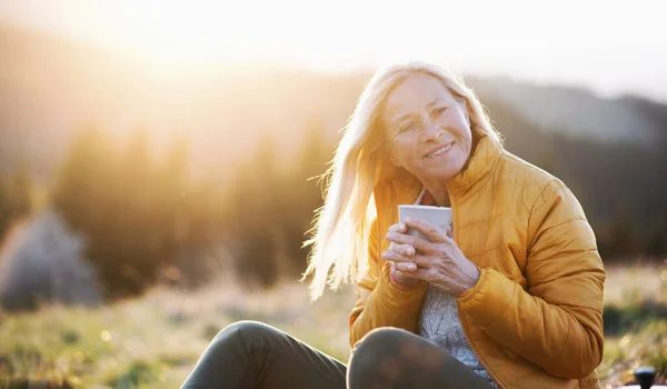 Atractiva mujer mayor sentada al aire libre en la naturaleza al atardecer, relajándose con café . — Foto de Stock