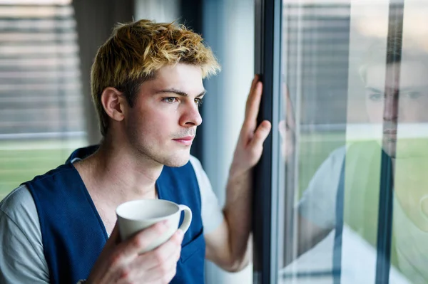 家でコーヒーを飲みながら窓から外を見る若い男. — ストック写真