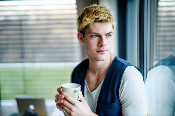 家でコーヒーを飲みながら窓から外を見る若い男. — ストック写真