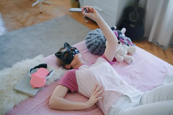 Jong meisje met kattenmasker op bed, selfie aan het nemen. Online dating concept. — Stockfoto