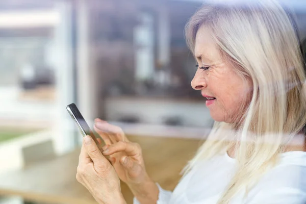 Porträtt av äldre kvinna som står inomhus, med hjälp av smartphone. — Stockfoto