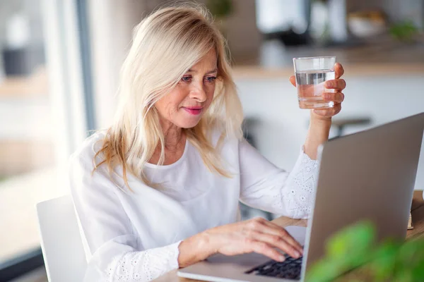 Senior vrouw met water en laptop binnen in het thuiskantoor, werken. — Stockfoto