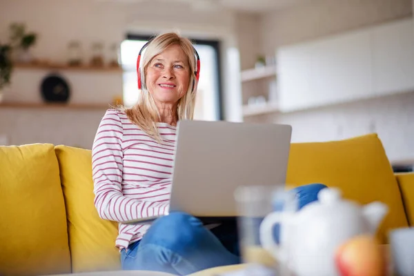 Oudere vrouw met koptelefoon en laptop binnen in het thuiskantoor, ontspannen. — Stockfoto