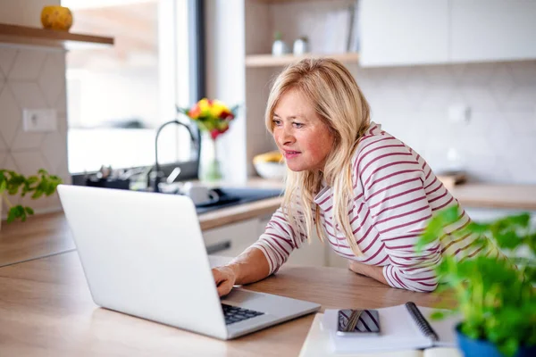 Senior vrouw met laptop binnen in keuken home office, werken. — Stockfoto