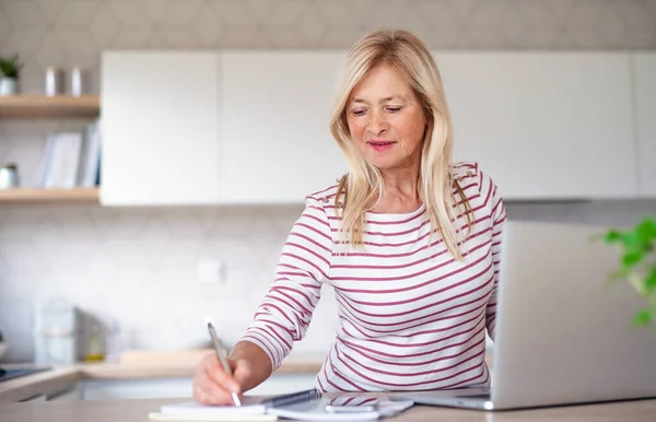 Senior kvinna med bärbar dator inomhus i köket hemmakontor, arbetar. — Stockfoto