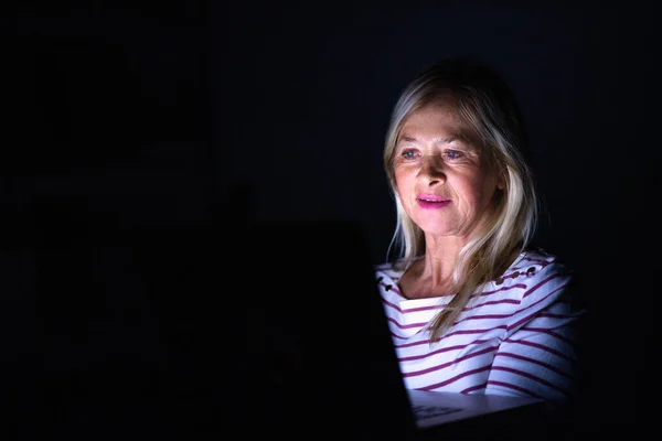 Senior woman with laptop indoors at home working late at night. — Stock Photo, Image