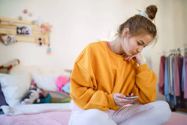 Giovane studentessa triste seduta sul letto, utilizzando lo smartphone . — Foto Stock
