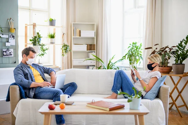 Casal com máscaras faciais sentado dentro de casa, usando a tecnologia . — Fotografia de Stock