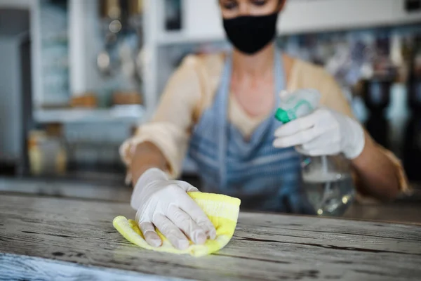 Proprietaria di caffetteria che lavora con maschera viso e guanti, disinfettante contatore . — Foto Stock