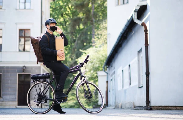 얼굴에 마스크를 쓰고 자전거를 타고 마을에서 스마트 폰을 사용하는 배달 남자. — 스톡 사진