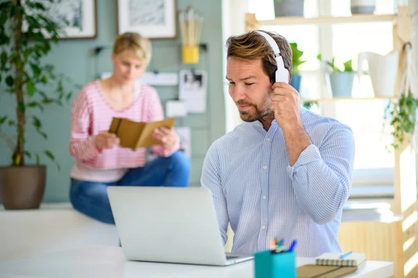 Gammal affärsman med hörlurar och bärbar dator inomhus i hemmakontoret, arbetar. — Stockfoto