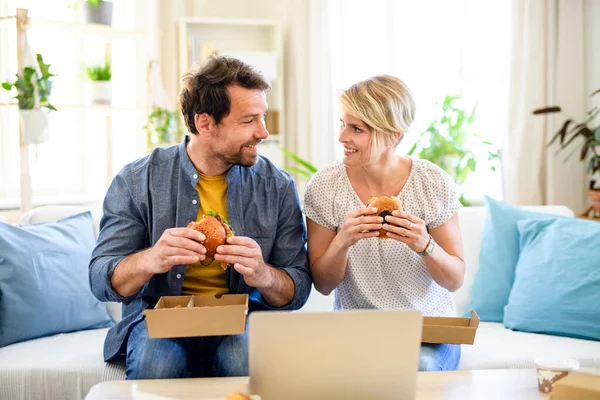 Happy couple sitting on sofa indoors at home, eating hamburgers. — Stock Photo, Image