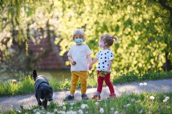 Małe dzieci z maskami twarzy i pies bawiący się na zewnątrz, koncepcja koronawirusa. — Zdjęcie stockowe
