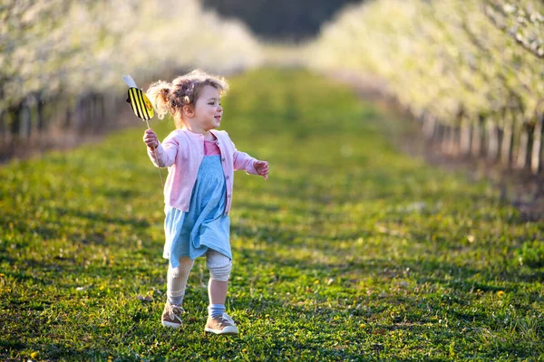 Piccola bambina che corre all'aperto nel frutteto in primavera, tenendo l'ape di carta . — Foto Stock