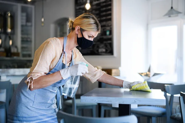 Propietaria de cafetería trabajando con mascarilla facial y guantes, desinfectando mesas . — Foto de Stock