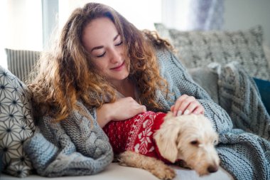 Young woman relaxing indoors on sofa at home with pet dog.
