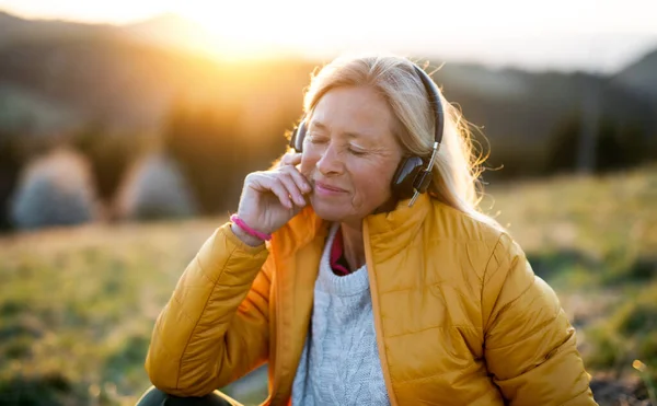Mujer mayor sentada al aire libre en la naturaleza al atardecer, relajándose con auriculares . — Foto de Stock