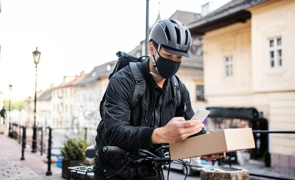 Entrega mensajero hombre con máscara facial y teléfono inteligente entrega caja de paquetes en la ciudad . — Foto de Stock