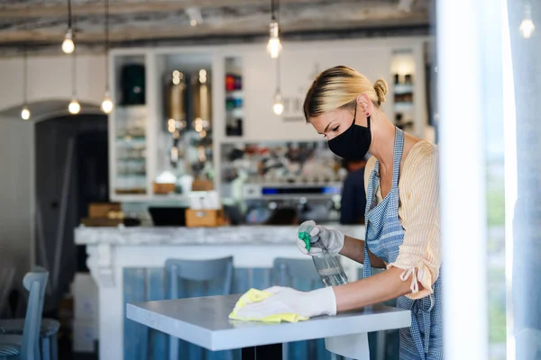 Kaffebutik kvinnlig ägare arbetar med ansiktsmask och handskar, desinficera bord. — Stockfoto