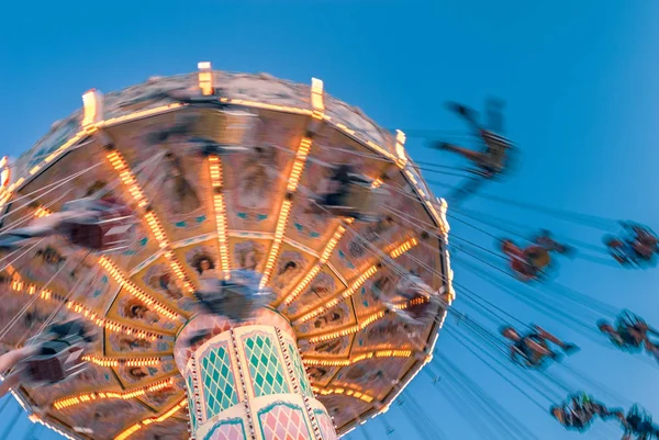 Amusement park motion blurred riders on retro vintage tilting swing against clear blue sky.