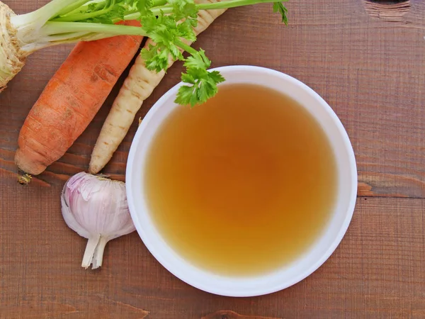 Caldo de ternera transparente, caldo de hueso, caldo en tazón blanco y verduras en vista superior de mesa de madera — Foto de Stock