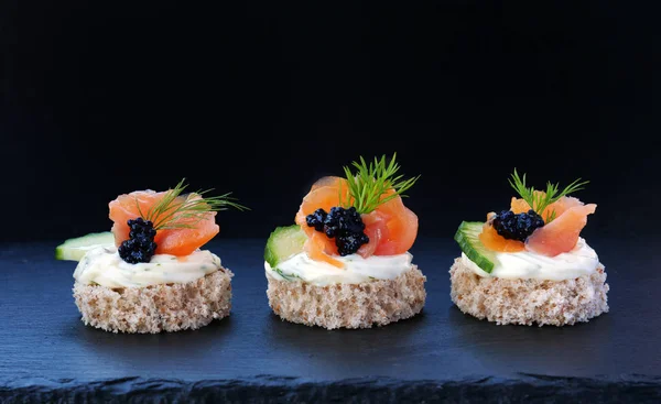 Canapés elegantes con salmón ahumado y caviar negro en empanadas de grano entero sobre bandeja de pizarra negra —  Fotos de Stock