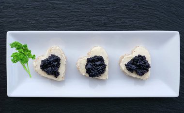 Kalp şeklinde parti ısırıkları, tereyağlı ekmek siyah bir havyar servis tepsisi üzerinde siyah levha, düz yatıyordu