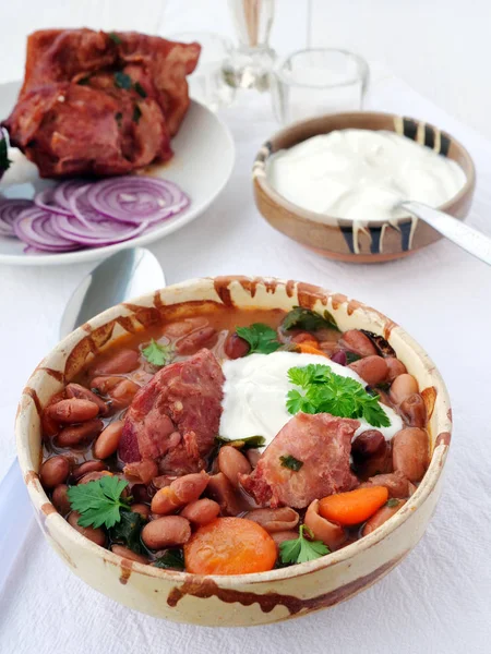 テーブルの上にサワークリームと赤玉ねぎとボウルにスモークハムホックとピント豆のスープ,垂直 — ストック写真